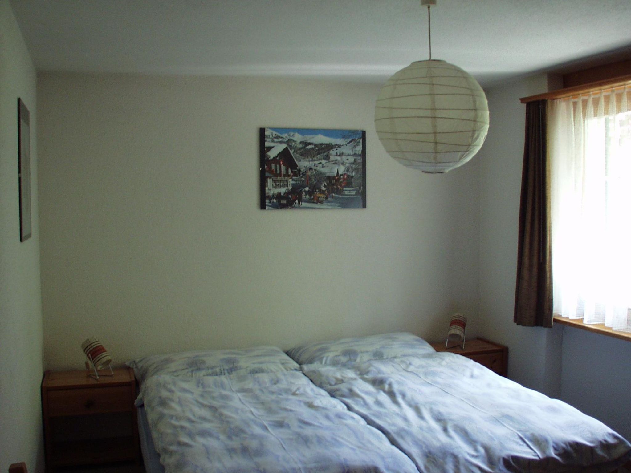 A bedroom with a bed, a nightstand, a lamp, a painting, and a window with curtains.