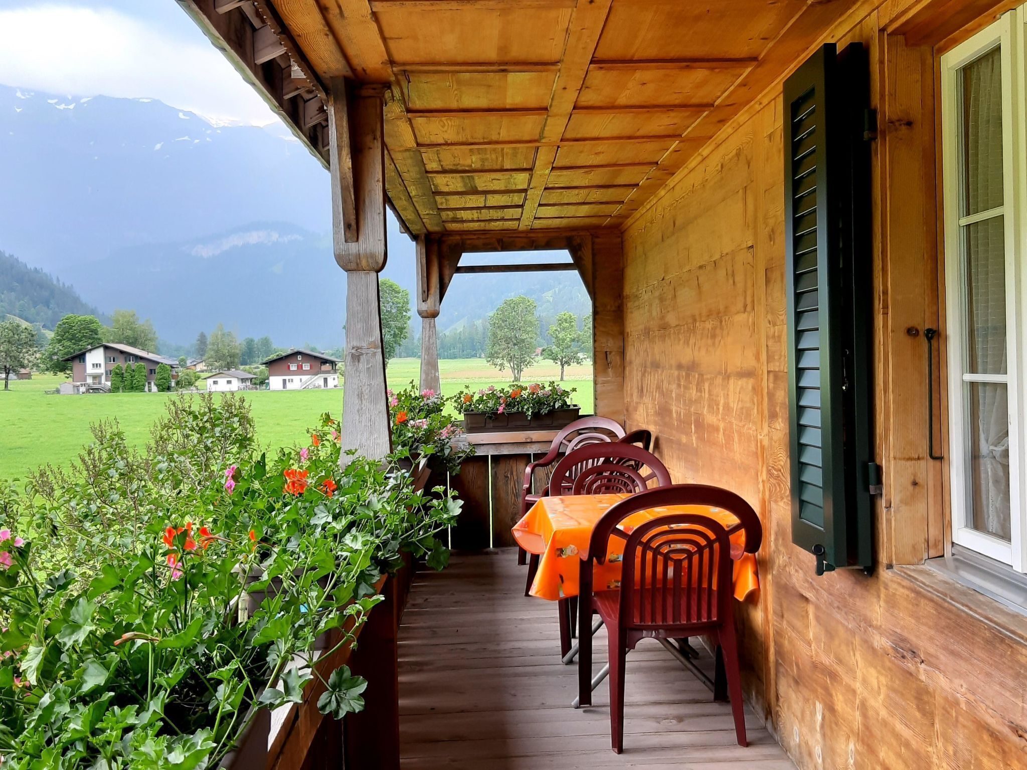 A wooden porch with a table and chairs, overlooking a green landscape and houses.