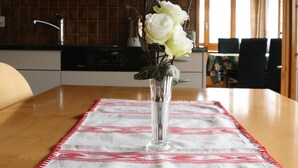 Une table à manger recouverte d’une nappe à motifs blancs et rouges, un vase en verre transparent contenant des fleurs blanches et un plafond en bois.
