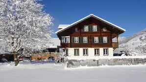 Un chalet en bois à deux étages, doté d’un toit en pente, entouré d’arbres couverts de neige et surplombant un ciel bleu dégagé.