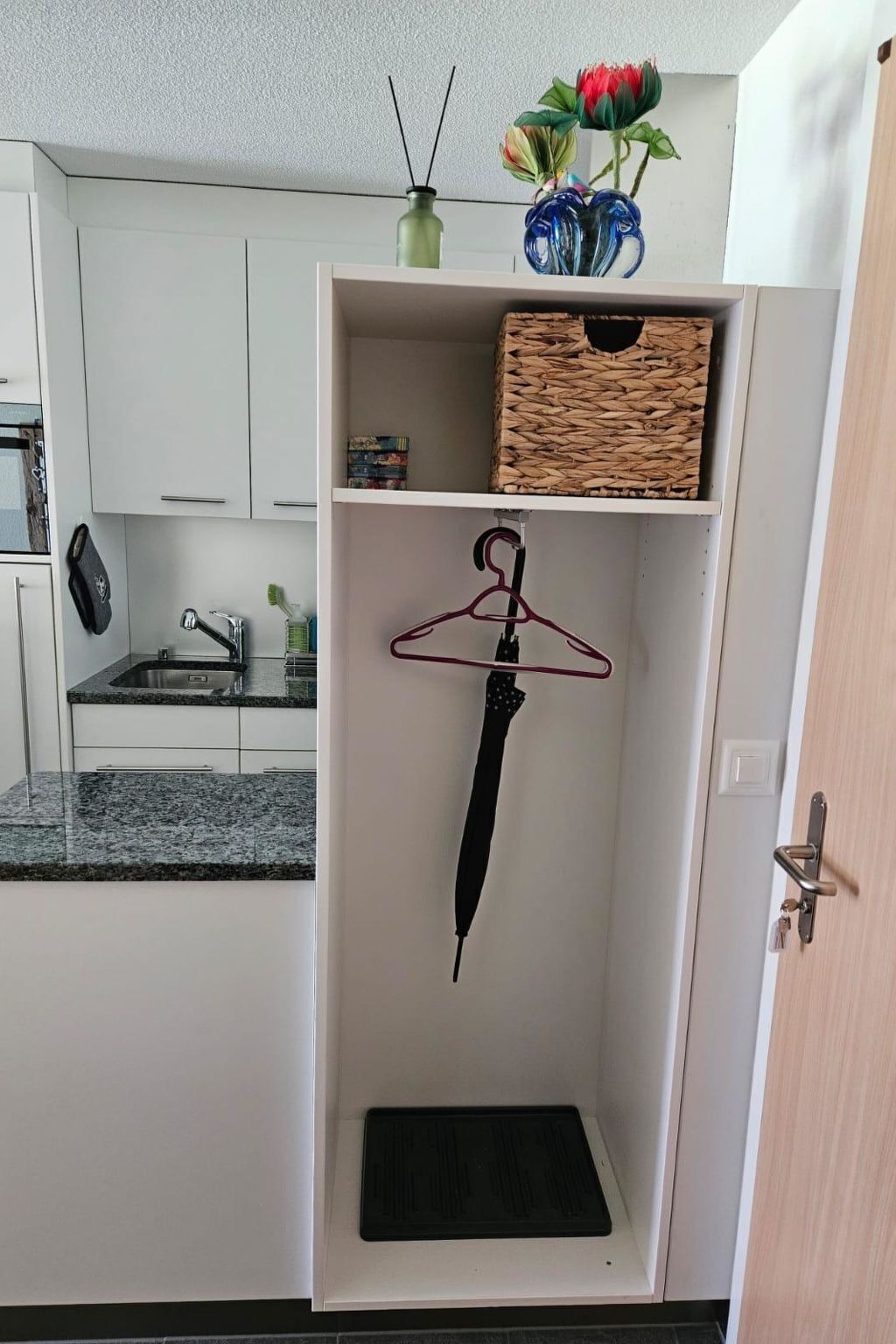 Uma cozinha com armários brancos, bancada de granito, um guarda-chuva preto pendurado e uma cesta de vime em uma prateleira.