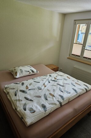 Ein Einzelbett mit gemustertem Überwurf, einem Kissen, einem Nachttisch und einem Fenster mit Jalousien.
