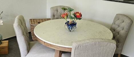 Une table de salle à manger ronde, entourée de quatre chaises, un vase rempli de fleurs, un luminaire suspendu et un coin cuisine avec des placards et un réfrigérateur en arrière-plan.