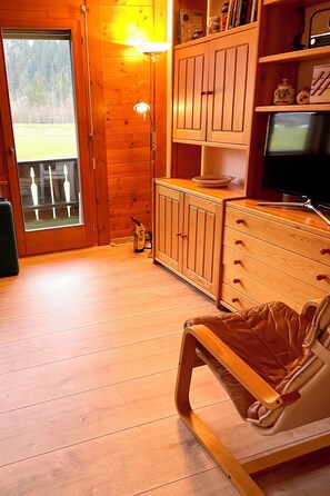 Ein Holzinterieur mit einer Schiebetür, die auf einen Balkon führt, einem Fernseher auf einem Holztisch und einem gepolsterten Sessel.