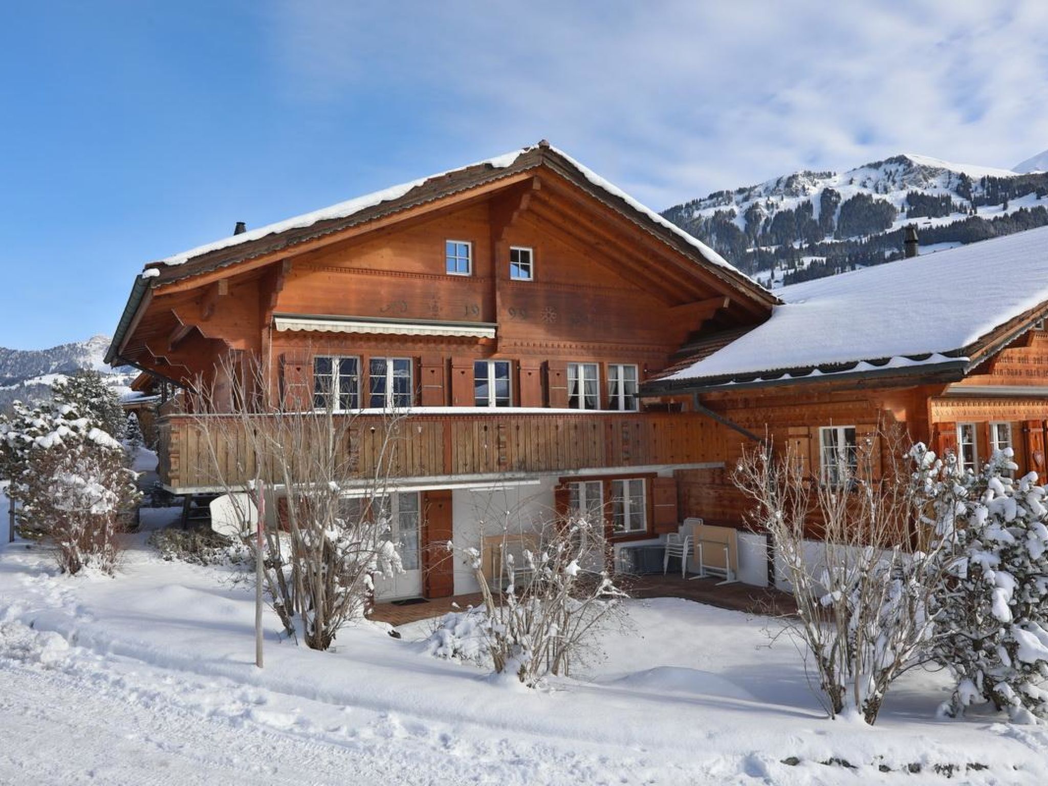 Ein großes Holzchalet mit Balkon, umgeben von verschneitem Gelände und Bäumen, mit Bergen im Hintergrund.