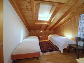 Una habitación pequeña de madera con dos camas, una mesita de noche, una silla y un tragaluz.