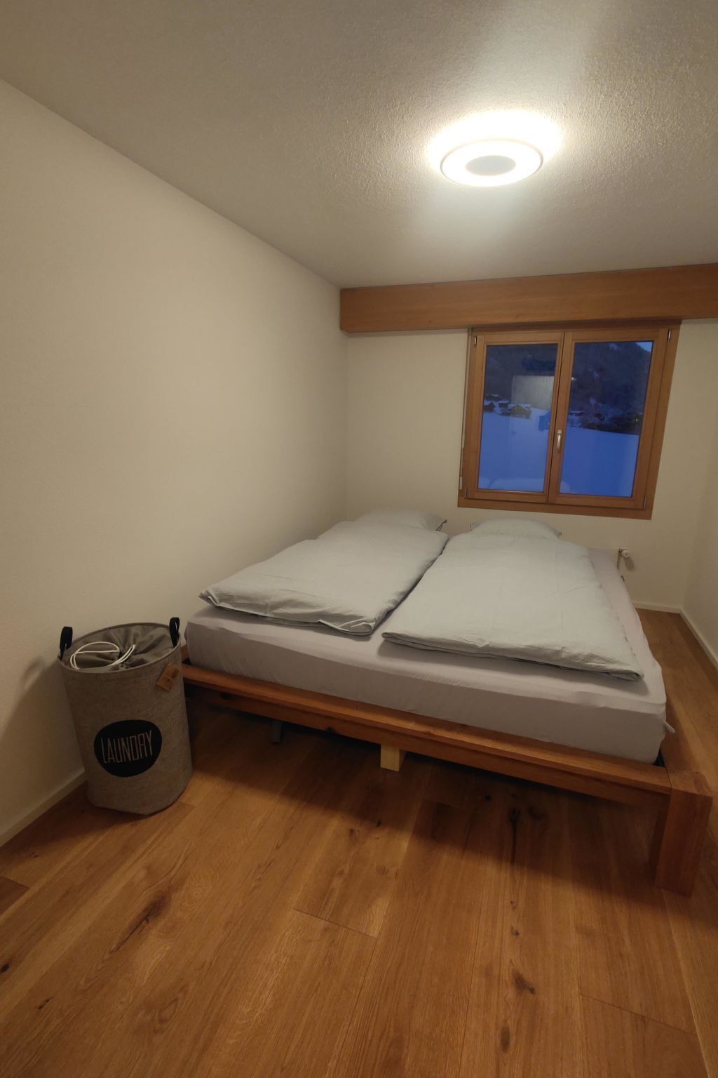 Ein Schlafzimmer mit einem hölzernen Bett, einem Fenster mit Blick auf eine verschneite Landschaft und einem Wäschekorb.