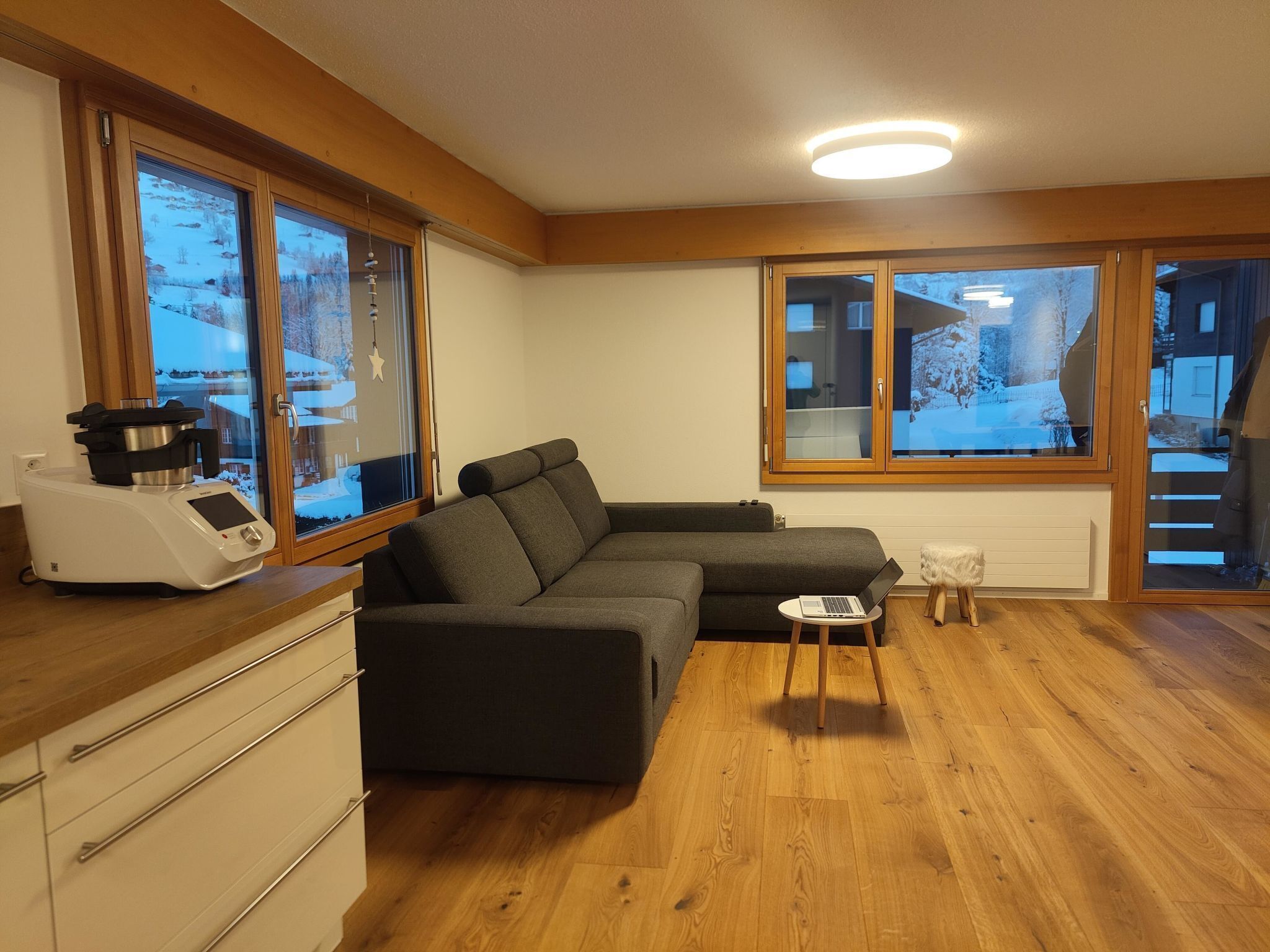 A modern living room with a sectional sofa, a small round table with a laptop, a built-in oven, and large windows with a view of a snowy landscape.
