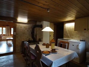 Ein Essbereich mit Tisch für zwei Personen, Holzstühle, einer Pendelleuchte und einer Küchenzeile mit weißen Geräten.