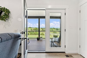 En åben dør, der fører ud til en balkon med udsigt over et grønt landskab og en klar himmel.