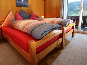 Ein hölzernes Bett mit roter Bettwäsche und mehreren Kissen, ein Holzboden und ein Blick auf eine gebirgige Landschaft durch ein großes Fenster.