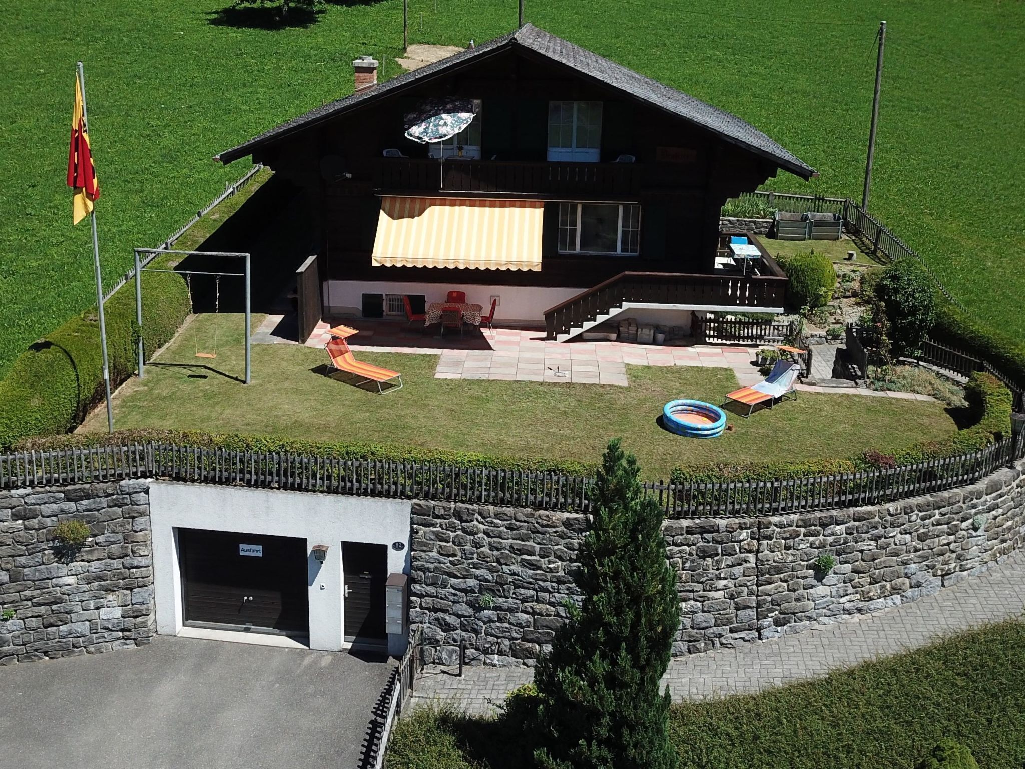 A two-story chalet with a stone wall entrance, a patio with outdoor furniture, and a swimming pool.