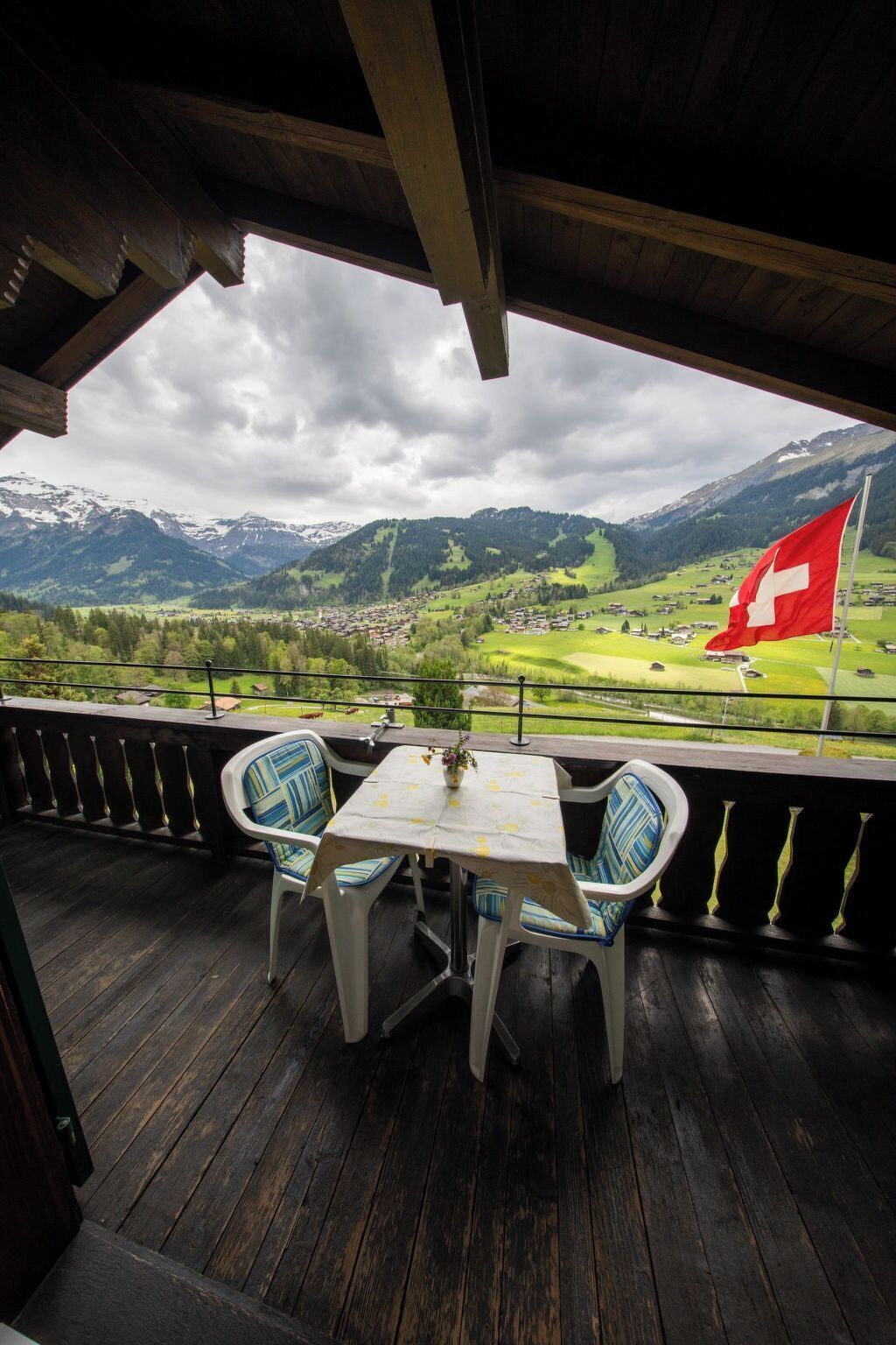 A balcony with a table and chairs, overlooking a village and mountains, with a Swiss flag visible.