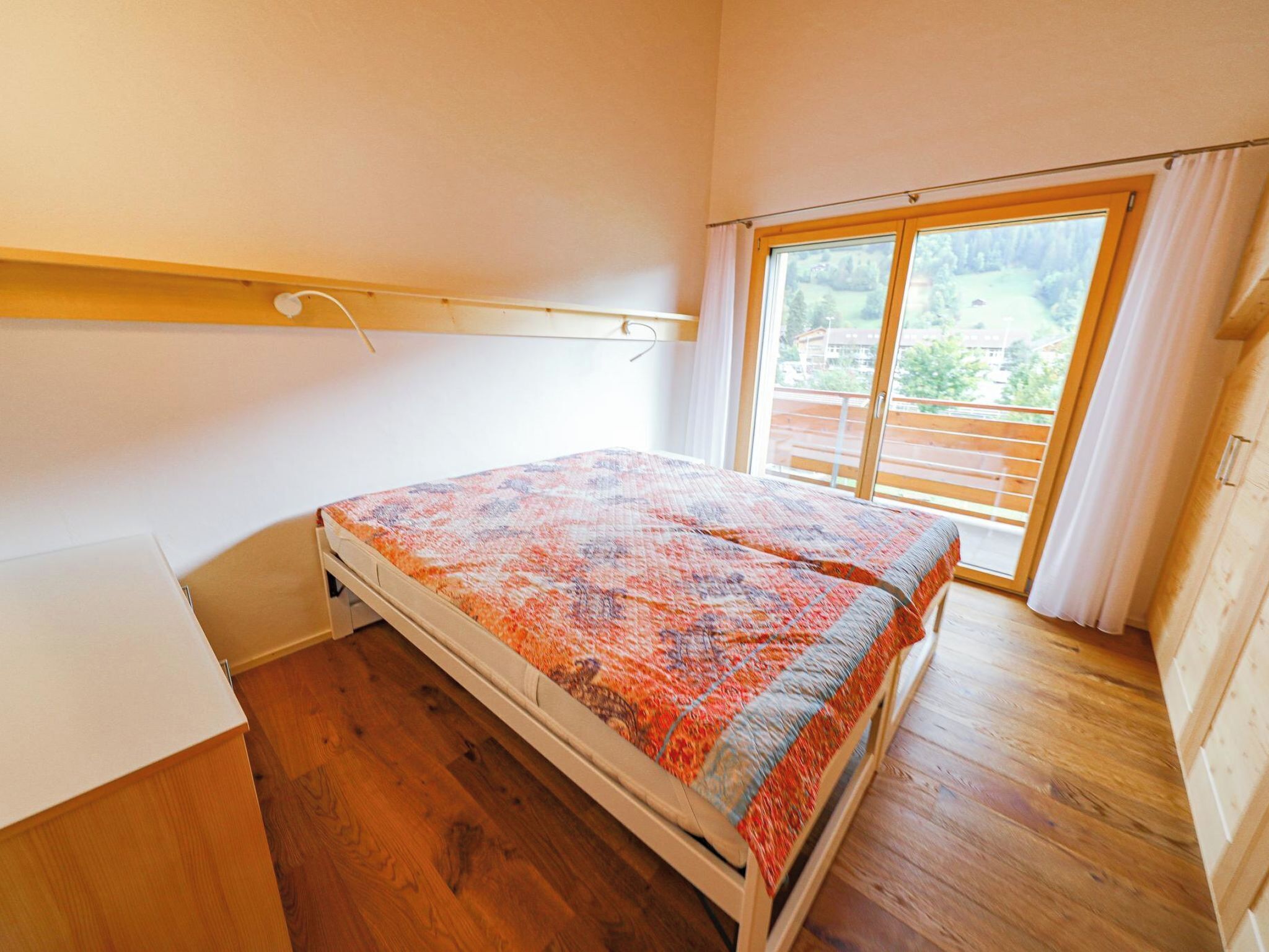 A bedroom with a bed, wooden flooring, a window with curtains, and a view of the outdoors.