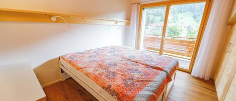 A bedroom with a bed, wooden flooring, a window with curtains, and a view of the outdoors.