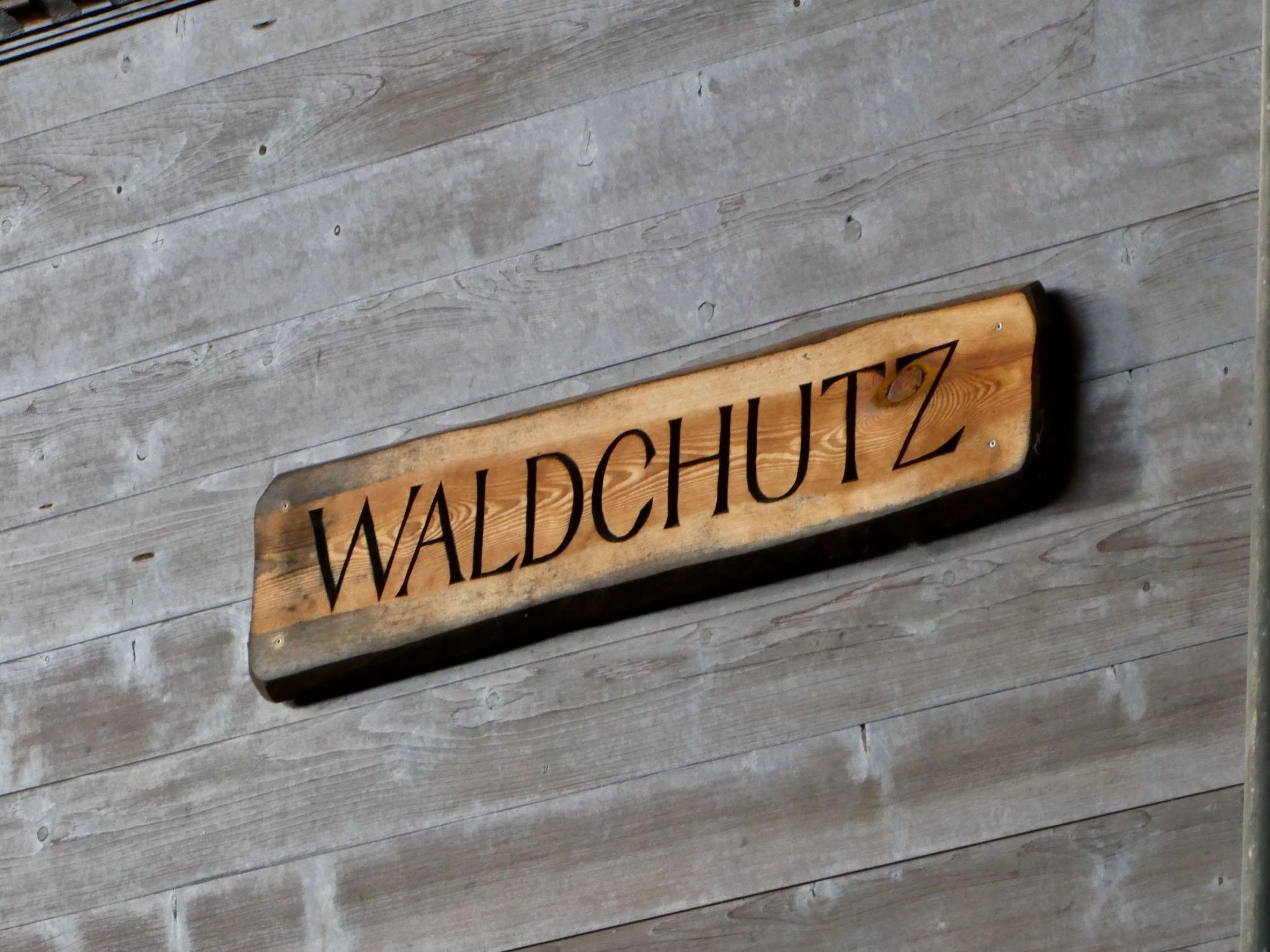 Een houten bord met de naam 'WALDCUTZ' bevestigd aan een muur met een houten planken achtergrond.