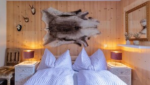 Ein Schlafzimmer mit großem Fellteppich, zwei Nachttischen mit Lampen, einem Bett mit gemusterten Kissen und Holzwänden mit aufgehängten Tierköpfen.