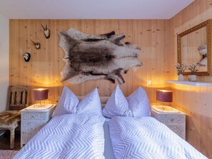Ein Schlafzimmer mit großem Fellteppich, zwei Nachttischen mit Lampen, einem Bett mit gemusterten Kissen und Holzwänden mit aufgehängten Tierköpfen.