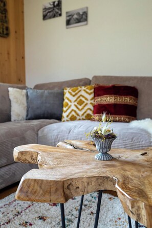 Ein Holztisch mit einem Blumenstrauß im Vordergrund, eine graue Couch mit gemusterten Kissen im Hintergrund und eine Holzvertäfelung an der Wand.