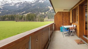 Ein Balkon mit Blick auf schneebedeckte Berge, eine Wiese und ein Holzinnenraum mit Tisch und Stühlen.
