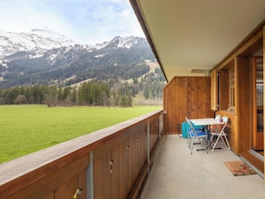 Ein Balkon mit Blick auf schneebedeckte Berge, eine Wiese und ein Holzinnenraum mit Tisch und Stühlen.