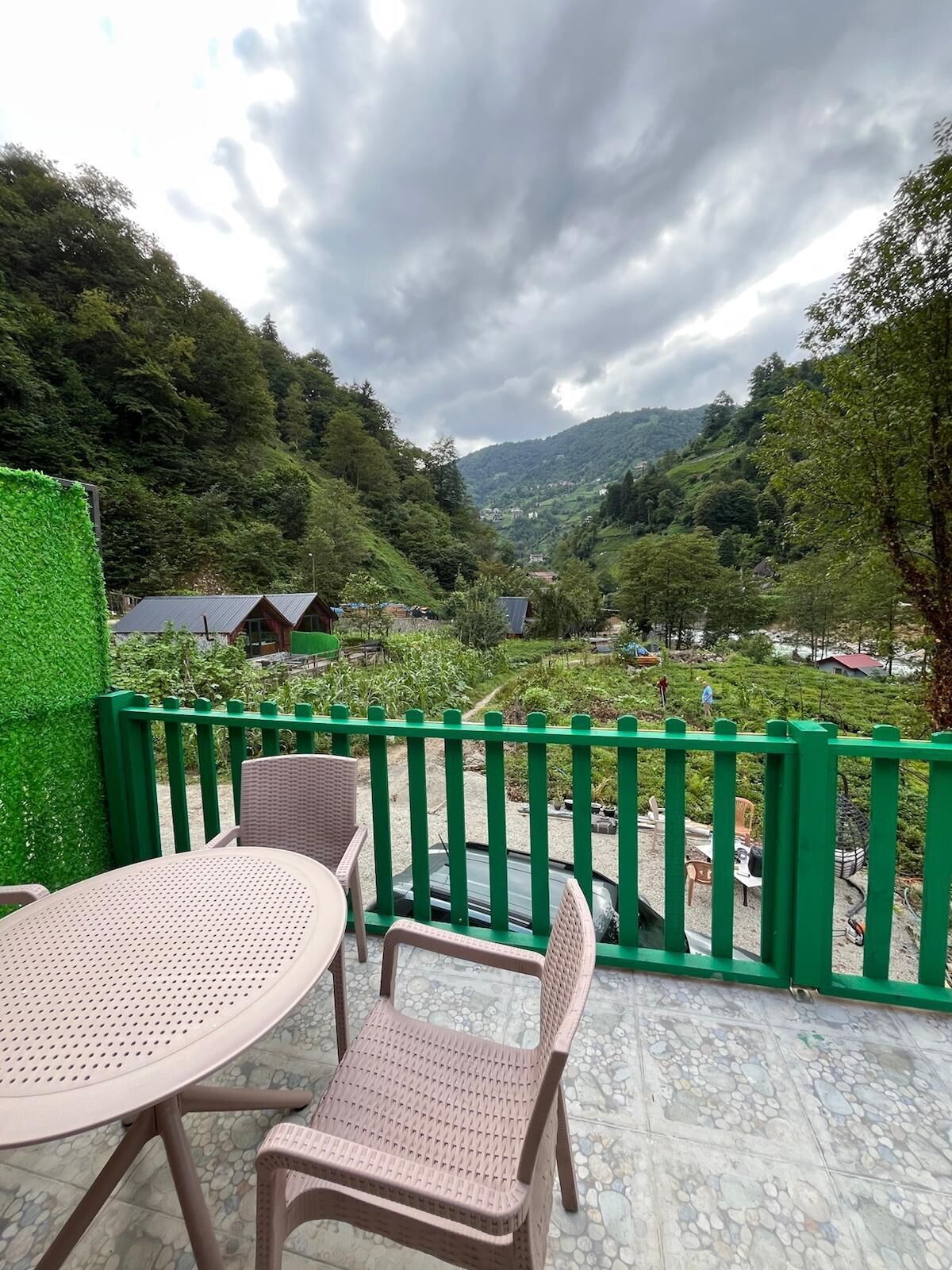 Una terraza con una mesa y sillas, barandal verde y vista a un valle con casas y árboles.