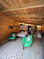 Interior de uma cabana de madeira com uma cama, duas poltronas verdes, um tapete pequeno e uma televisão.