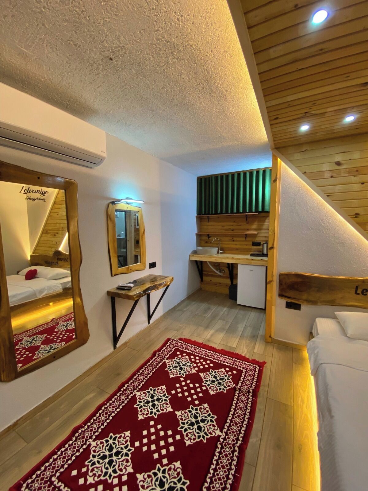 A hotel room with a wooden ceiling, a bed with white bedding, a mirror, a small table, and a red patterned rug.
