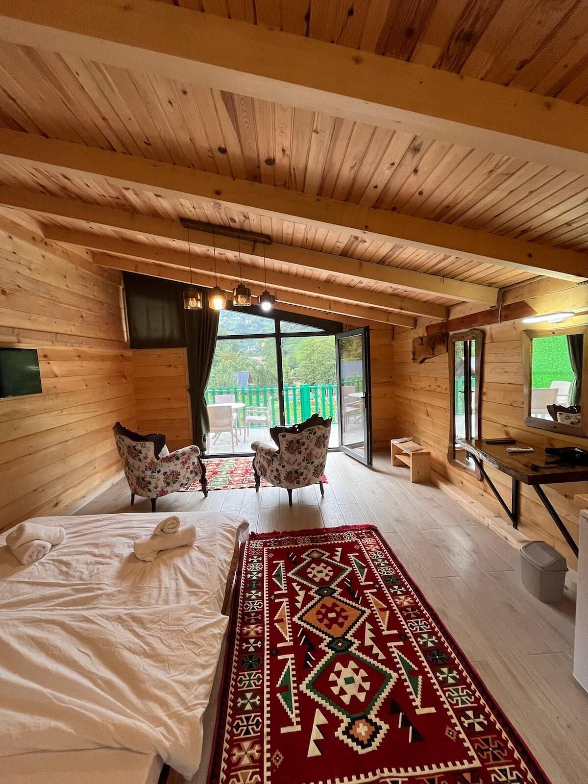 A wooden cabin room with a bed, two chairs, a desk, and a large window.