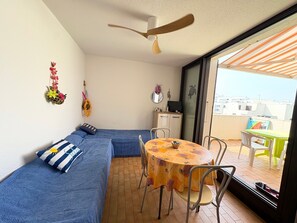 A room with a blue sofa, a round table with chairs, a ceiling fan, and a view through a glass door. - T2 apartment sea view - 4 guests (Le Barcarès)