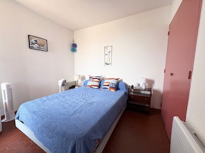 A bedroom with a bed, a bedside table, a lamp, a heater, and a wall with a framed picture. - T2 apartment sea view - 4 guests (Le Barcarès)