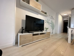 Ein modernes Wohnzimmer mit einem Flachbildfernseher auf einer weißen Konsole, Holzboden und einer Wandablage mit dekorativen Gegenständen.