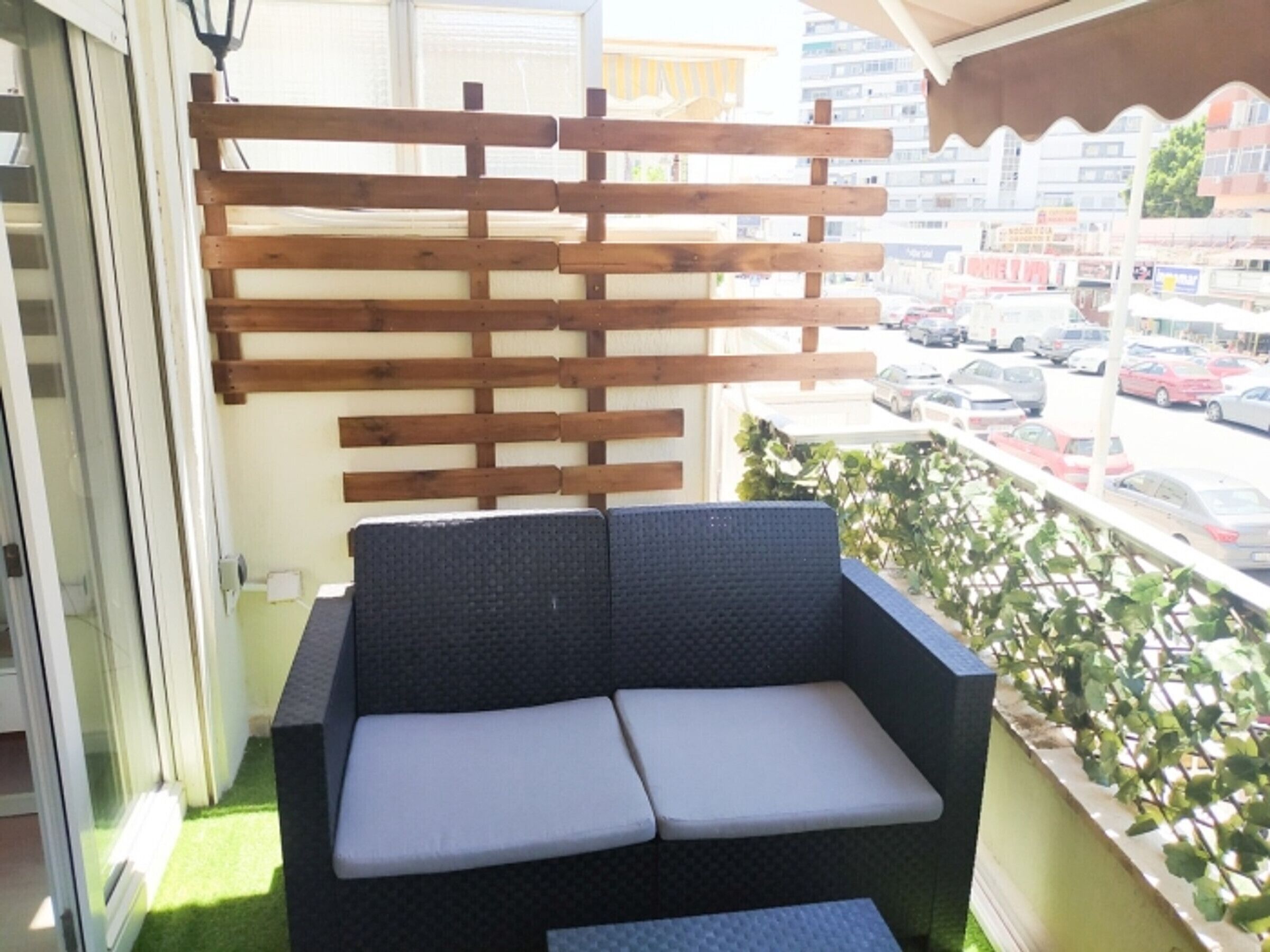 Un balcón con una barandilla de madera, un sofá tejido negro con cojines blancos y vista a un estacionamiento.