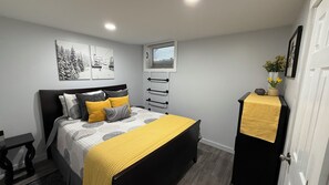 Een slaapkamer met een bed, nachtkastje, dressoir, raam met jaloezieën en wanddecoratie.