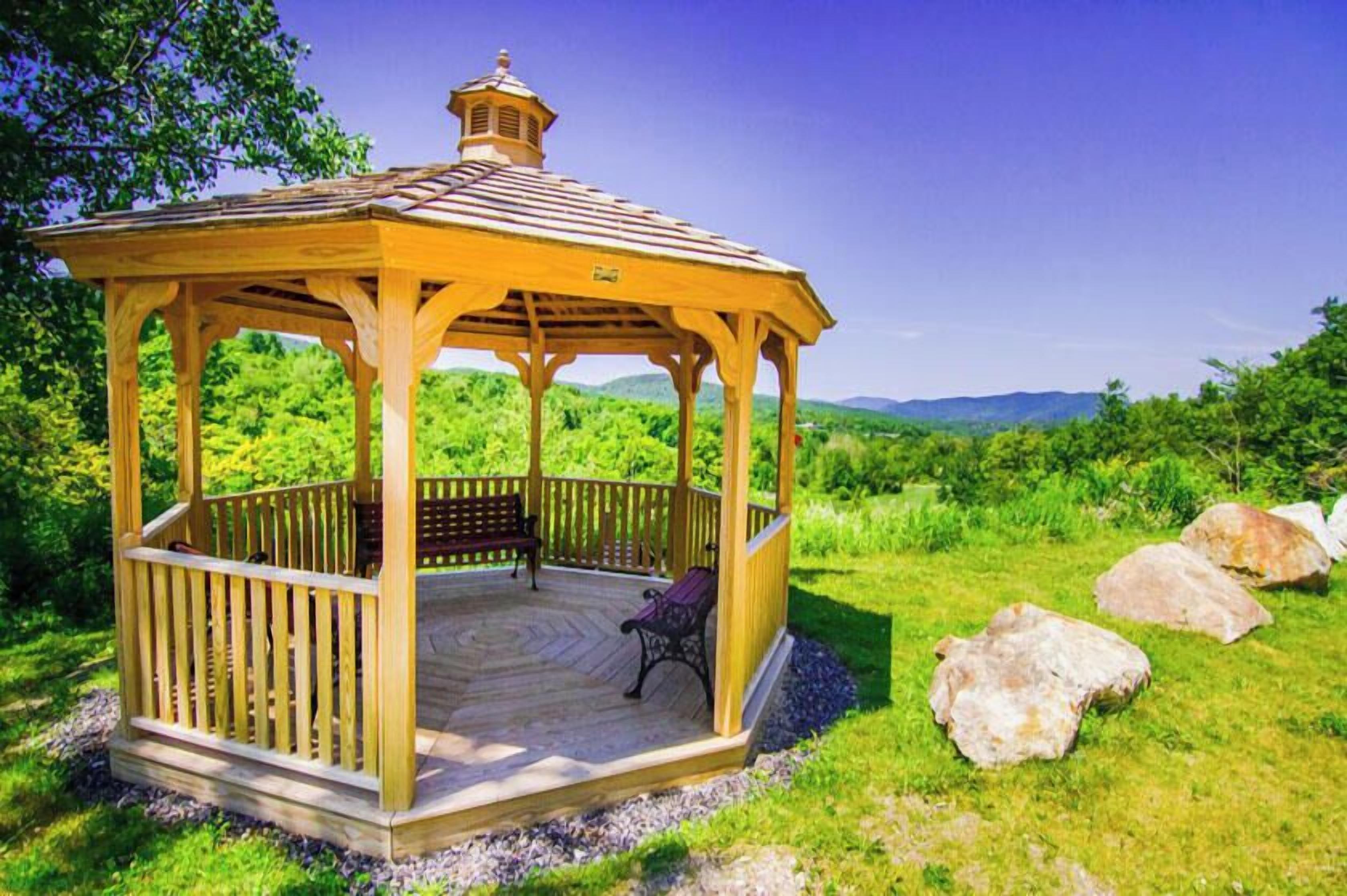 Un kiosque en bois avec un banc et une chaise, entouré d’herbe et de rochers.