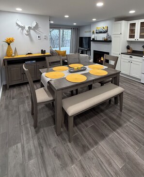 Een moderne eethoek met een houten tafel en bank, een open haard en een keuken op de achtergrond.