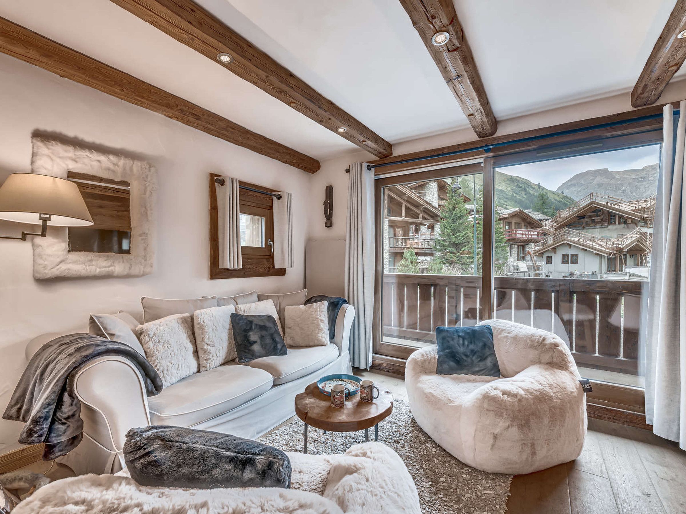 Un salon chaleureux avec un canapé, un fauteuil et un pouf, une grande fenêtre donnant sur un village de montagne, des poutres en bois au plafond et une lampe murale.