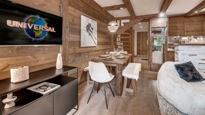 A modern interior with a wooden ceiling, a dining area, a sofa, and a television displaying 'UNIVERSAL'. - Apartment with parking - 6 guests (Val d'Isère)