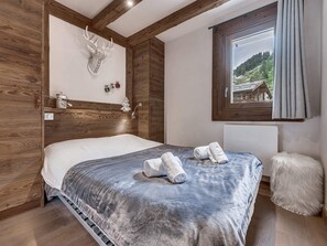 A bedroom with a wooden headboard, a large bed with a grey blanket, two rolled towels, a white fluffy stool, and a window with a view of a mountain landscape. - Apartment with parking - 6 guests (Val d'Isère)