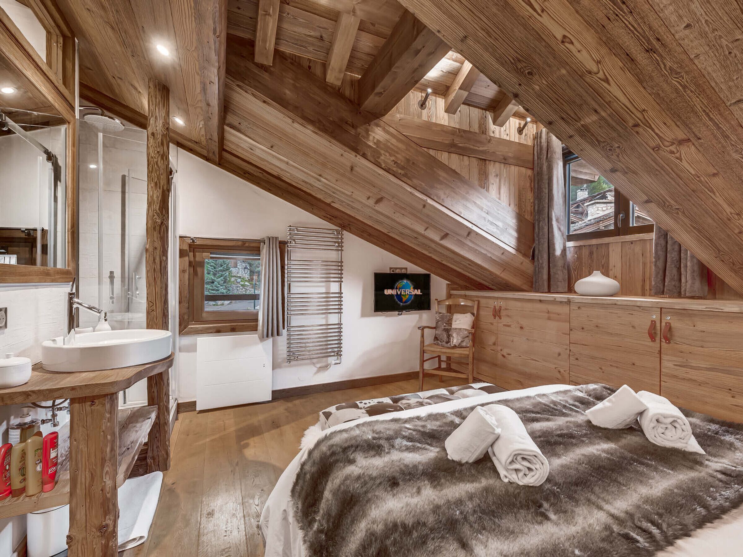 A bedroom with a sloped ceiling, wooden beams, a large bed with a fur throw, a wooden cabinet, and a television on the wall.