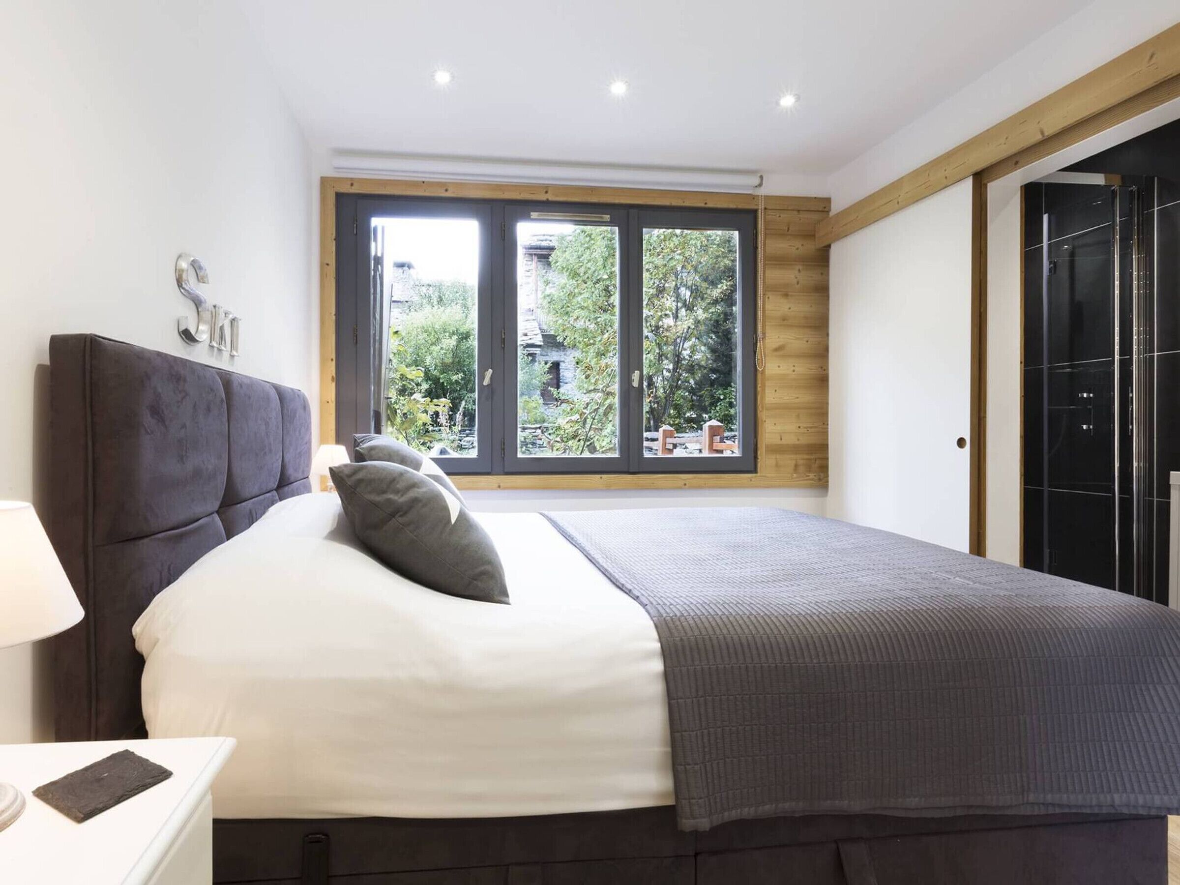 A modern bedroom with a large bed, a nightstand, a window with a view of greenery, and a wooden ceiling.