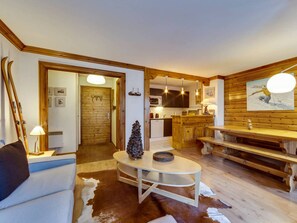 A cozy living room with a wooden floor, a white sofa, a coffee table, and a fireplace. There is a kitchen area with wooden cabinets and a dining table. - Comfortable apartment at ski slopes - 6 guests (Val-d'Isère)