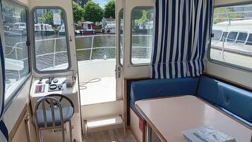 Et bådinteriør med kontrolpanel, et lille bord, en bænk og udsigt til vandet samt fortøjede både.