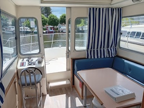 A boat interior with a control panel, a small table, a bench, and a view of the water and docked boats.