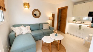 A modern living room with a sofa, two small tables, a kitchen area with cabinets, and a door leading to another room. - Beach Getaway with a Pool and Parking (AN)