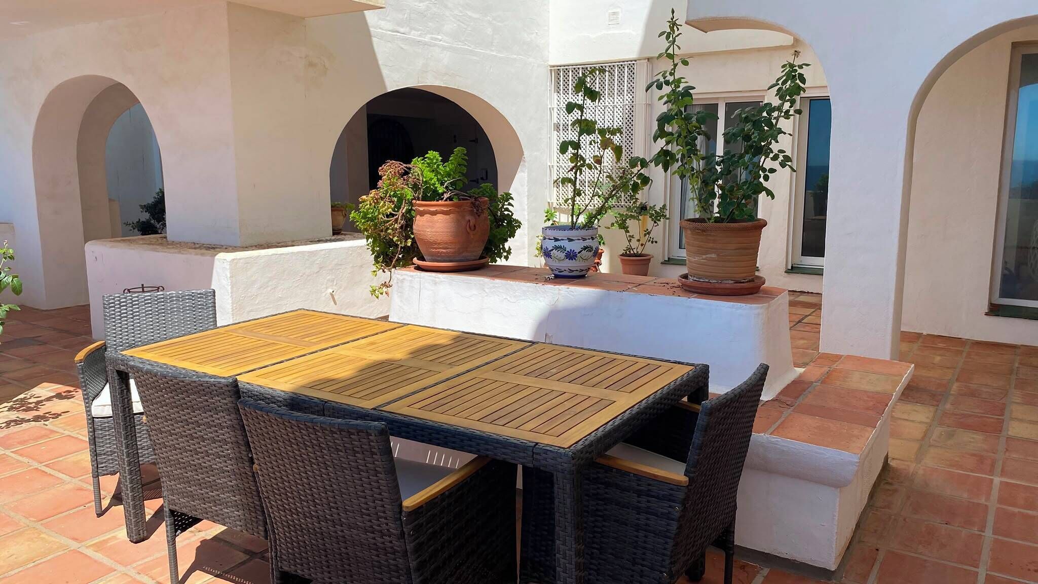 Een terras buiten met een houten tafel en rieten stoelen, omgeven door potplanten en witte muren.