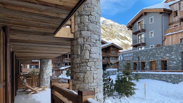 Ein Hotel mit Stein- und Holzarchitektur, einer überdachten Außenbereits und einem verschneiten Berg im Hintergrund.