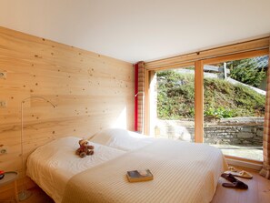 Ein Schlafzimmer mit Bett, einem Teddybären, einem Buch, Hausschuhen und einem Fenster mit Blick auf Grünflächen.