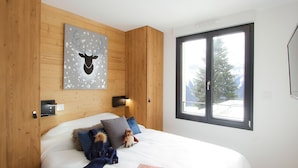 Une chambre à coucher avec un lit, des oreillers, une couverture, un jouet en peluche, un livre, une œuvre d’art accrochée au mur, une fenêtre et une porte.