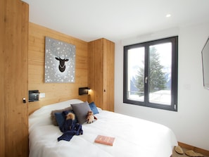 Ein Schlafzimmer mit einem Bett, Kissen, einer Decke, einem Stofftier, einem Buch, einem Wandbild und einem Fenster sowie einer Tür.